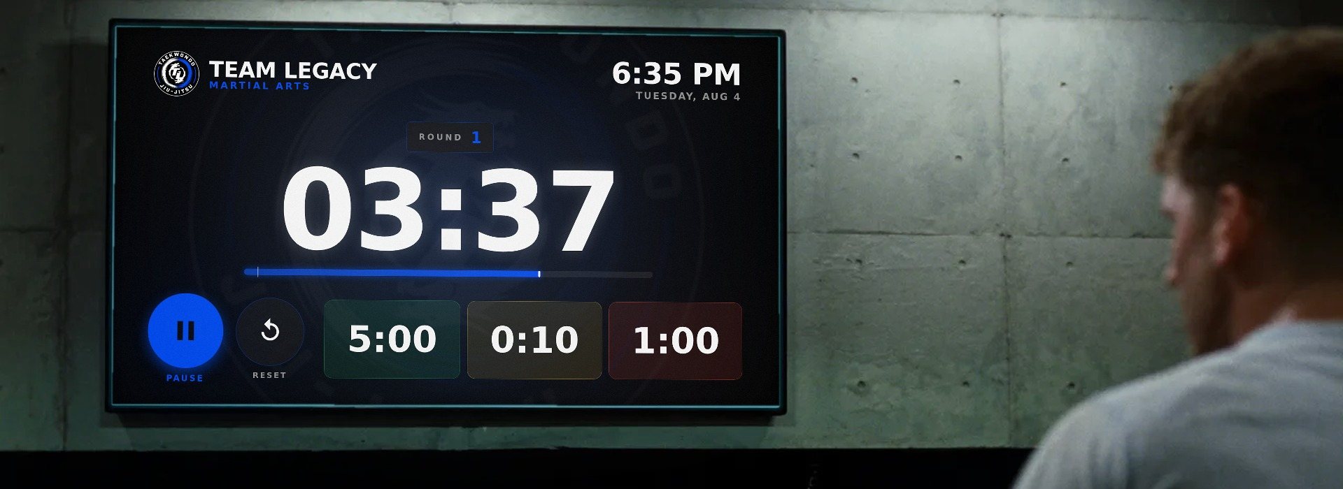 A round timer on a screen mounted on the concrete wall of a gym, carrying Team Legacy's name and colors with three minutes and thirty-seven seconds left in round one, and a fighter in the foreground turned to look at it